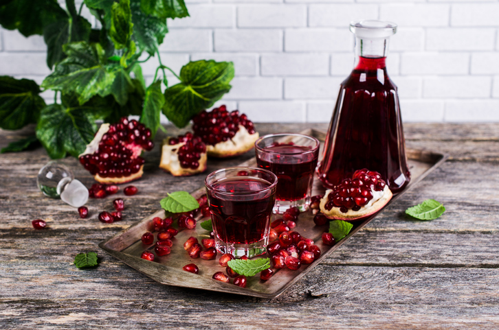 Auf einem rustikalen Holztablett stehen zwei Gläser mit dunkelrotem Likör neben einer passenden Glaskaraffe, dekoriert mit frischen Minzblättern und Granatapfelkernen.