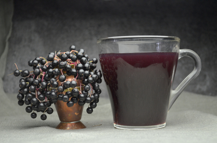 Ein Glas dunkler Holundersaft steht neben einer dekorativen Vase mit frischen, schwarzen Holunderbeeren auf grauem Leinen.