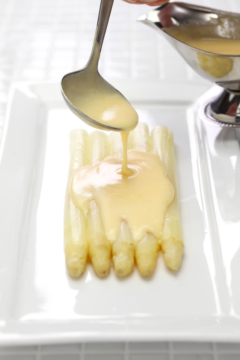Sauce Hollandaise wird mit einem Löffel über weißen Spargel auf einem Teller gegeben.