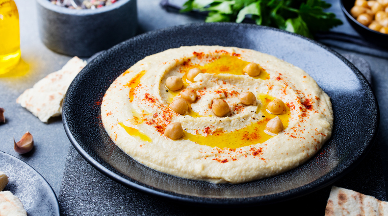 Kichererbsen-Hummus ist auf einem dunklen Teller mit Olivenöl, Paprikapulver und ganzen Kichererbsen angerichtet.