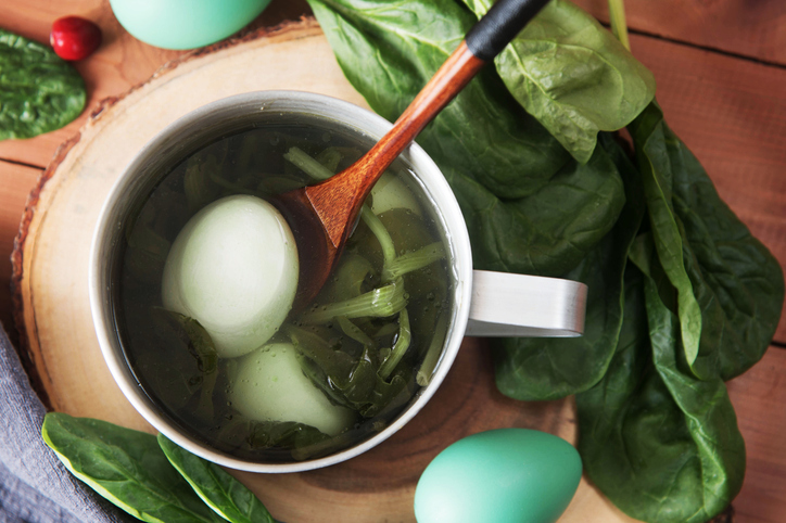 Zwei hellgrün gefärbte Eier liegen in einer Metalltasse mit Wasser und frischen Spinatblättern auf einer Holzscheibe.