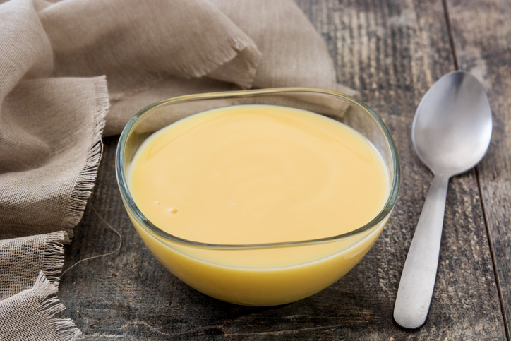Eine kleine Glasschale mit cremigem Vanillepudding steht auf einem rustikalen Holztisch neben einem Löffel und einem hellen Stofftuch.