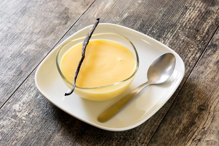 Eine kleine Glasschale mit Vanillepudding auf einem weißen Teller, dekoriert mit einer Vanilleschote und einem Löffel auf einem Holztisch.