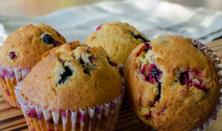 Vier goldbraun gebackene Muffins mit dunklen Johannisbeeren in gemusterten Papierförmchen auf einem Holzbrett.
