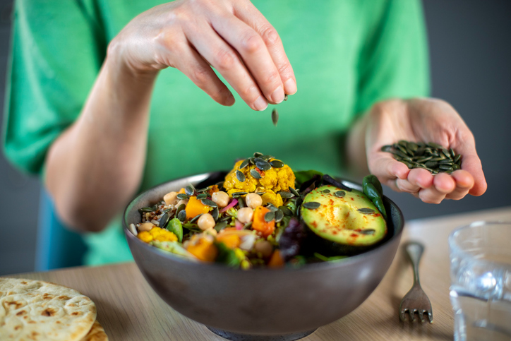 Jemand streut eine Handvoll grüne Kürbiskerne über eine bunte Bowl mit geröstetem Blumenkohl, Avocado und Kichererbsen.