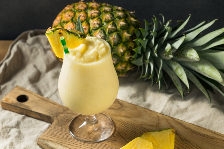 Ein bauchiges Glas gefüllt mit gefrorener Pina Colada auf einem Holzbrett, im Hintergrund eine ganze Ananas.