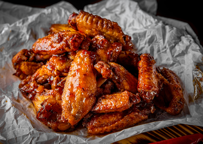 Ein großer Berg glänzender Chicken Wings liegt auf zerknittertem weißem Einschlagpapier, daneben eine rote Chilischote.