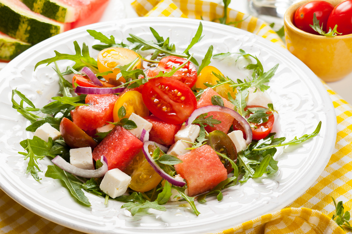 Ein weißer Teller mit einem frischen Salat aus Wassermelonenstücken, gelben und roten Tomaten, Feta-Würfeln und Rucola.