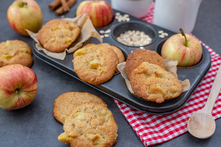 Nahaufnahme von selbstgebackenen Apfel-Hafer-Cookies neben frischen Äpfeln und einem Holzlöffel auf einem dunklen Hintergrund.