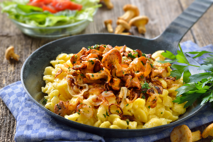 Eine Eisenpfanne mit Eierspätzle, Röstzwiebeln und knusprig gebratenen Pfifferlingen auf einem blau-weiß karierten Küchentuch.