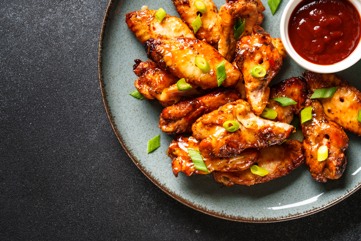Draufsicht auf einen Teller mit glasierten Chicken Wings und einer kleinen weißen Schale voller dunkelroter Chilisauce.