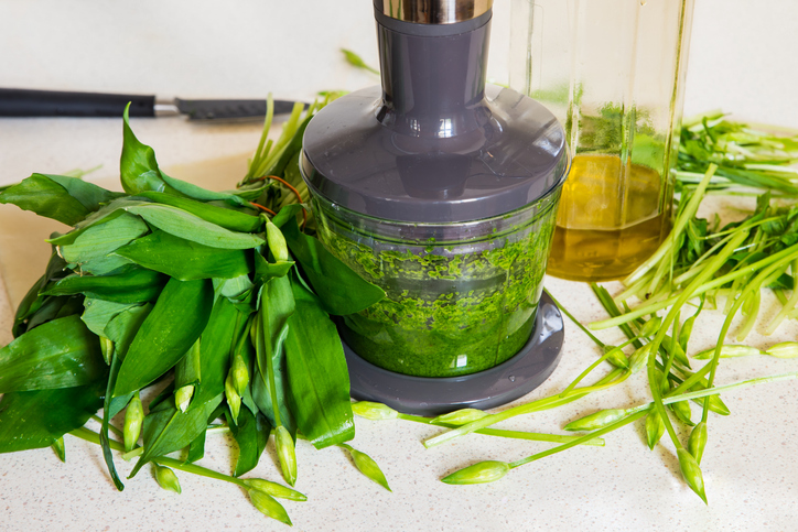 Ein großer Bund frischer Bärlauch liegt neben einem Mixer mit grüner Pesto-Masse.