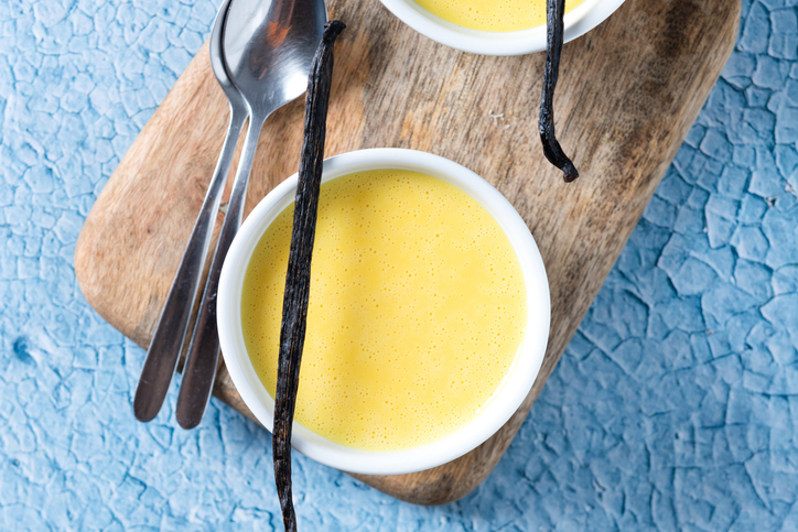 Ein einzelnes Förmchen mit Vanillepudding steht auf einem Holzbrett, daneben liegen zwei Löffel und eine lange Vanilleschote.