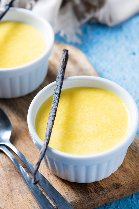 Zwei weiße Dessertförmchen mit feinem, gelbem Vanillepudding stehen auf einem rustikalen Holzbrett, garniert mit einer Vanilleschote.