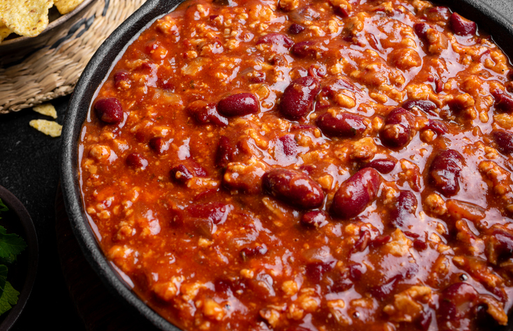 Ein Nahaufnahme eines würzigen Chilis in einer dunklen Pfanne, in dem Kidneybohnen in einer sämigen Tomatensauce mit Hackfleisch zu sehen sind.