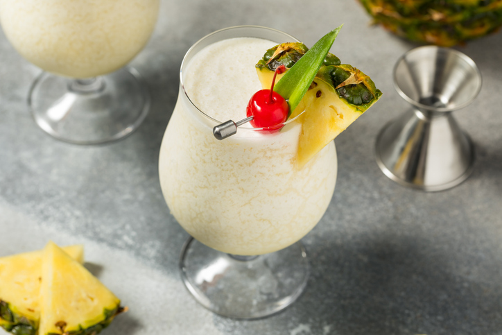 Eine Pina Colada in einem eleganten Kelchglas, garniert mit Ananasstücken, grünen Blättern und einer leuchtend roten Kirsche.