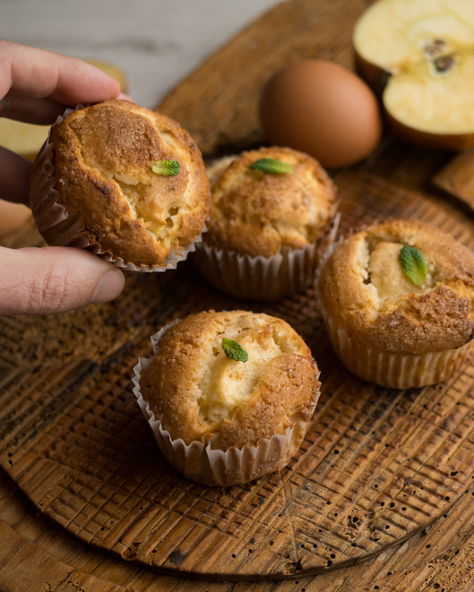 Eine Hand nimmt einen Apfel-Zimt-Muffin von einem rustikalen Holzbrett, weitere Muffins stehen daneben, dekoriert mit kleinen Minzblättern.