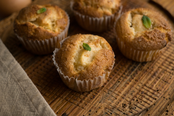 Mehrere Apfel-Zimt-Muffins mit einem kleinen Minzblatt-Topping auf einem strukturierten Holzbrett, daneben ein beiges Leinentuch.