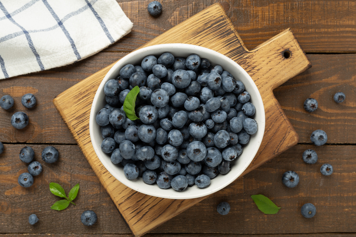 Eine weiße Schüssel randvoll mit frischen Blaubeeren steht auf einem rustikalen Holzbrett, umgeben von einzelnen Beeren und Blättern.