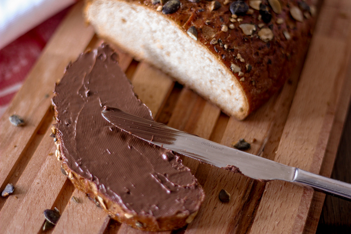 Eine Scheibe dunkles Körnerbrot auf einem Holzbrett, die gerade großzügig mit cremiger Schokoladencreme bestrichen wurde.