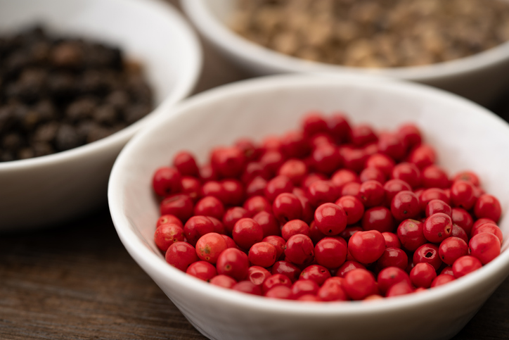 Eine weiße Schale gefüllt mit leuchtend roten rosa Pfefferkörnern steht neben weiteren Schalen mit anderen Gewürzen auf einem dunklen Holztisch.