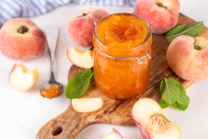 Ein Glas gefüllt mit leuchtend orangefarbener Marmelade auf einem Holzbrett, umgeben von frischen Pfirsichen und Minzblättern.