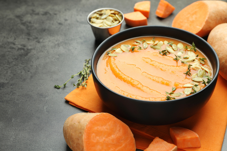 Eine Hand streut frische Kürbiskerne auf eine Portion orangefarbene Süßkartoffelsuppe in einer dunklen Schale.