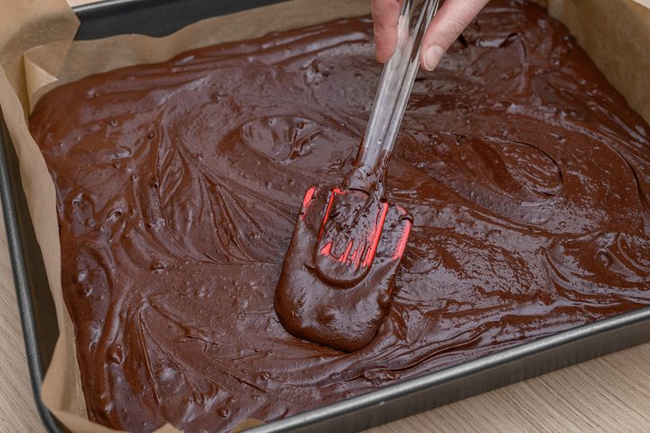 Eine Hand streicht mit einem roten Teigschaber die dunkle Brownie-Masse gleichmäßig bis in die Ecken der Backform glatt.