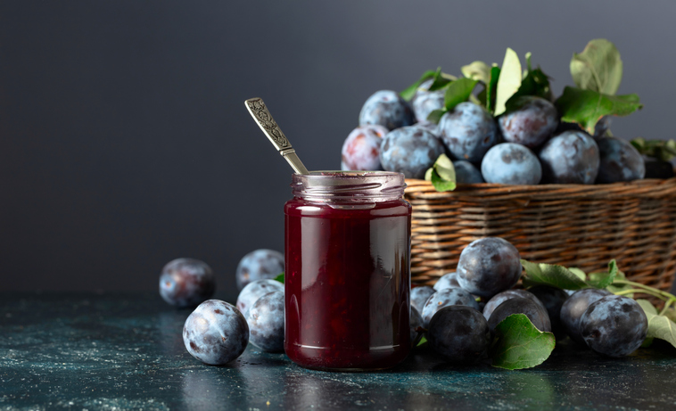 Ein Glas dunkles Pflaumenmus mit einem silbernen Löffel steht neben einem rustikalen Weidenkorb voller blauer Früchte.