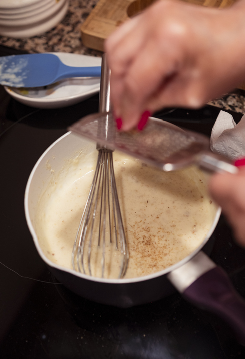 Eine Person reibt frische Muskatnuss über eine cremige, helle Béchamelsauce in einem Topf, während ein Schneebesen darin steht.