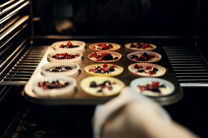 Ein Muffinblech mit zwölf gefüllten Förmchen, die reichlich mit roten und schwarzen Beeren belegt sind, wird in einen dunklen Backofen geschoben.