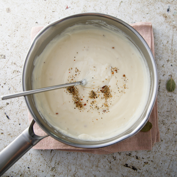Eine frisch zubereitete, glatte Béchamelsauce in einem Edelstahltopf auf einem Stofftuch, bestreut mit frisch gemahlenem Pfeffer.