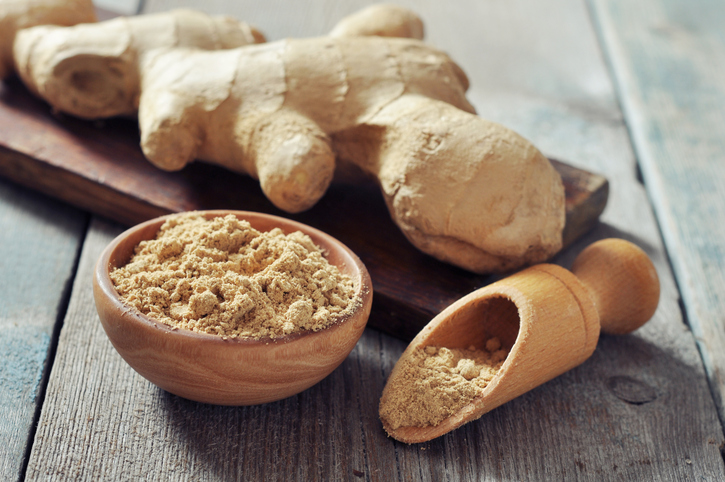 Eine große frische Ingwerknolle liegt neben einer kleinen Holzschale mit fein gemahlenem Ingwerpulver und einer hölzernen Schaufel auf einem rustikalen Holztisch.