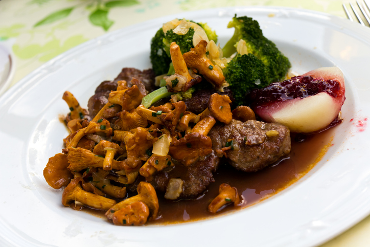 Ein weißer Teller mit gebratenem Fleisch, Brokkoli, Preiselbeeren und einer reichhaltigen Portion Pfifferlinge in Sauce.