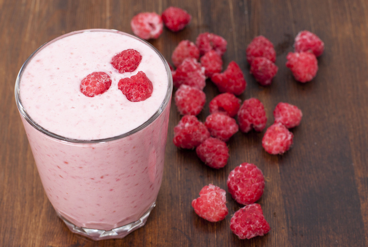 Ein Glas rosa Himbeer-Shake auf dunklem Holz, dekoriert mit drei Himbeeren auf der Schaumkrone.