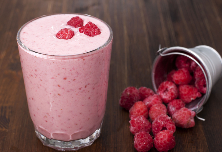 Ein Glas cremiger Himbeer-Shake neben einem kleinen Metalleimer, aus dem frische Himbeeren auf einen dunklen Holztisch kullern.