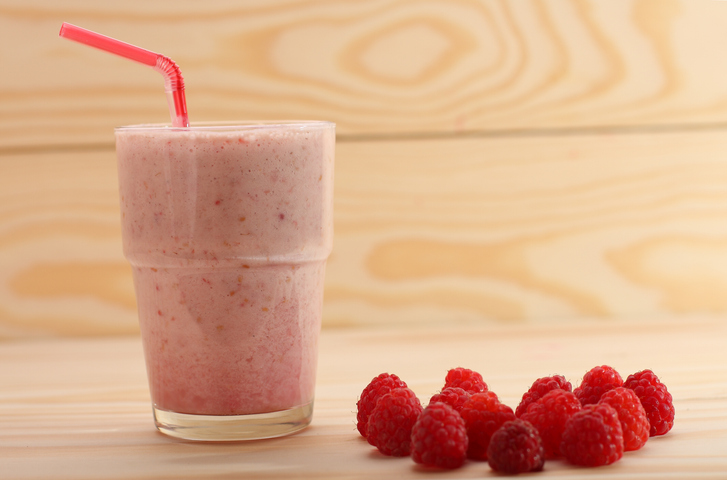 Ein einfaches Glas Himbeer-Joghurt-Shake mit rotem Trinkhalm vor einer hellen Holzwand, davor liegen einige frische Himbeeren.