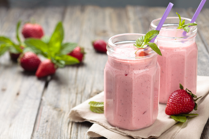 Zwei Gläser mit einem cremigen rosa Erdbeershake, garniert mit frischer Minze und Erdbeeren auf einem rustikalen Holztisch.