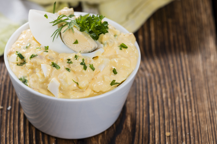 Eine weiße Schüssel gefüllt mit cremigem Eiersalat, garniert mit einem hartgekochten Ei und frischer Petersilie auf Holzgrund.