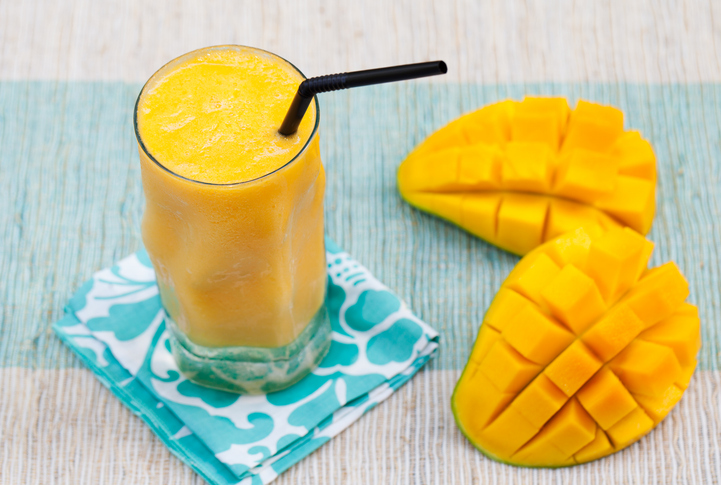 Ein erfrischender Mangoshake in einem Glas mit schwarzem Strohhalm, begleitet von zwei aufgeschnittenen Mangohälften auf einem bunten Tuch