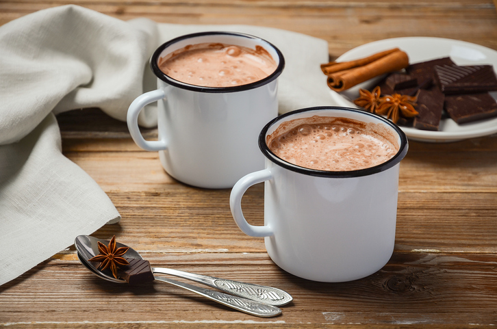 Zwei weiße Emaille-Becher gefüllt mit schaumiger heißer Schokolade auf einem rustikalen Holztisch, daneben Zimtstangen, Sternanis und Schokoladenstücke.