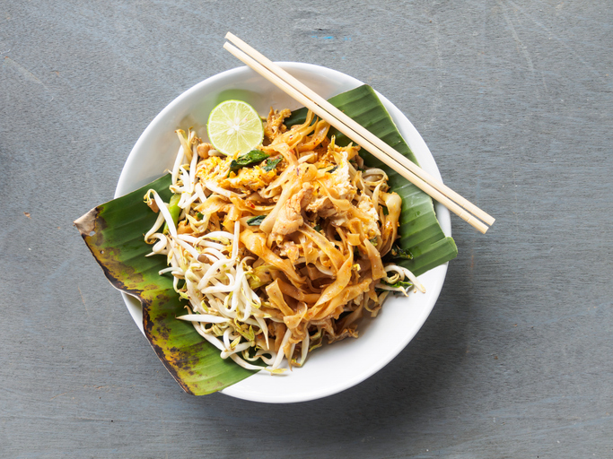 Eine Portion Pad Thai in einer weißen Schale auf einem Bananenblatt, dazu Stäbchen und eine Limettenscheibe