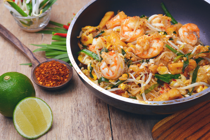 Pad Thai mit großen Garnelen, Sojasprossen und Kräutern in einer Pfanne auf einem Holztisch, serviert mit Limette und Chilipulver