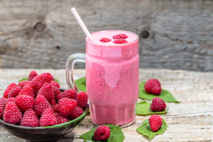 Ein großes Glas Himbeer-Joghurt-Shake mit einem Trinkhalm neben einer vollen Schale frischer Himbeeren.
