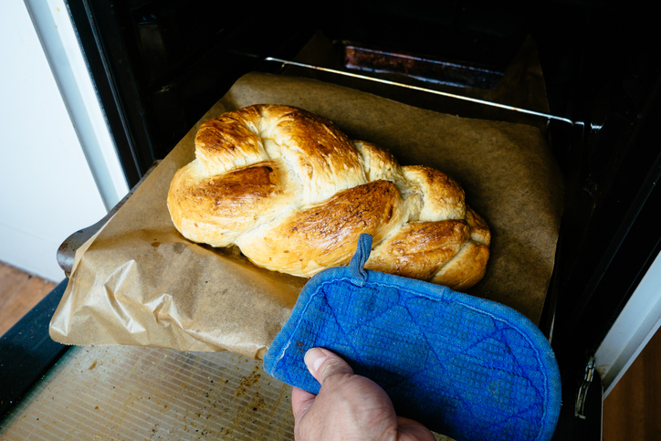 Eine Hand mit blauem Topflappen zieht ein Backblech mit einem frisch gebackenen Hefezopf aus dem Ofen.
