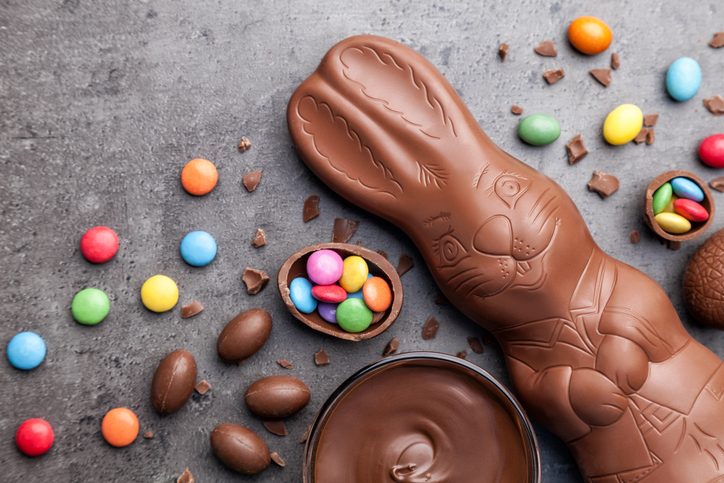 Ein großer Schokoladen-Osterhase liegt neben bunten Schokolinsen und kleinen gefüllten Schokoeiern auf einem dunklen Steingrund.
