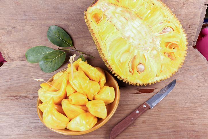 Eine halbierte frische Jackfrucht liegt neben einer Schale mit verzehrfertigen, gelben Fruchtfleischstücken und einem Messer auf Holz.