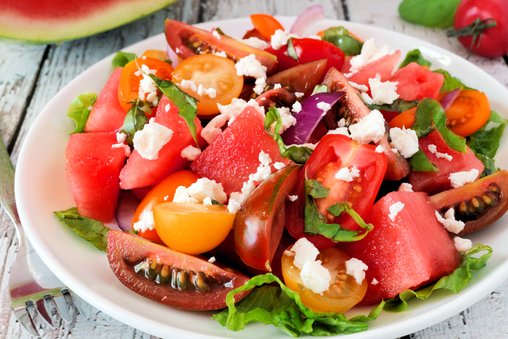Nahaufnahme eines Salats mit großen Wassermelonenstücken, Tomatenhälften, roten Zwiebeln und zerbröseltem Feta, garniert mit frischem Basilikum.