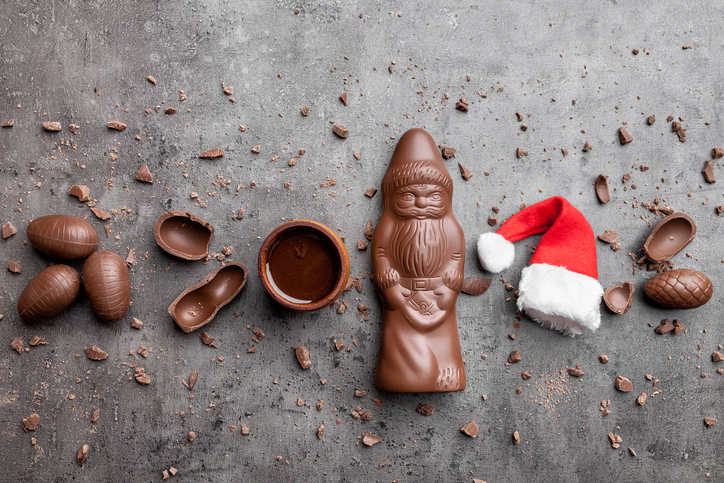 Ein Schokoladen-Nikolaus auf grauem Untergrund, umgeben von Schokoladeneiern, einer kleinen Weihnachtsmütze und Schokosplittern.