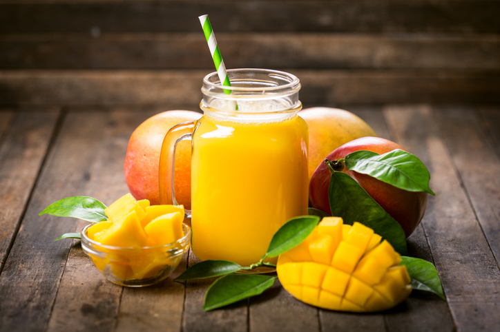 Ein leckerer Mangoshake in einem Mason Jar mit einem gestreiften Strohhalm, umgeben von frischen Mango-Stücken und Blättern auf einem rustikalen Holztisch